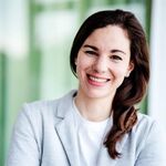 Lächelnde Frau mit braunen Haaren, in einem grauen Blazer, vor einem grünen Hintergrund.
