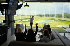 Ein Golfer schwingt einen Schläger, während Zuschauer aus einer Lounge-Bereich an einer Driving Range jubeln.