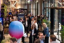 A busy conference scene with attendees networking and a mascot in a colorful outfit.